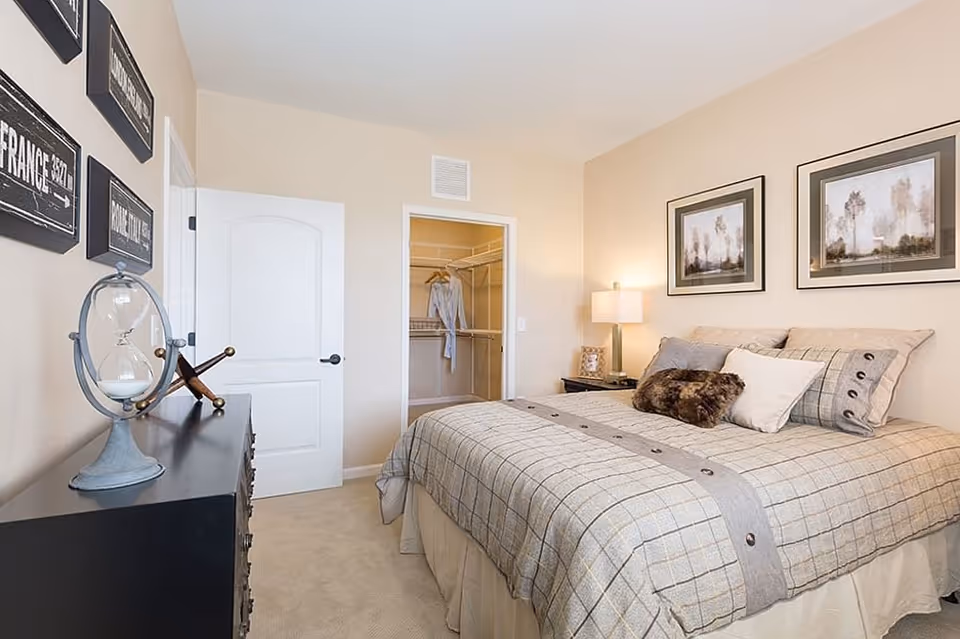 A neatly made bed with a plaid bedspread and multiple pillows in a bedroom. There are two framed pictures on the wall above the bed. A nightstand with a lamp and a small decorative item is next to the bed. Across from the bed is a black dresser with a decorative hourglass and a small sculpture on top. The door to a walk-in closet is open, showing hanging clothes inside. The walls are painted beige and the carpet is light-colored.