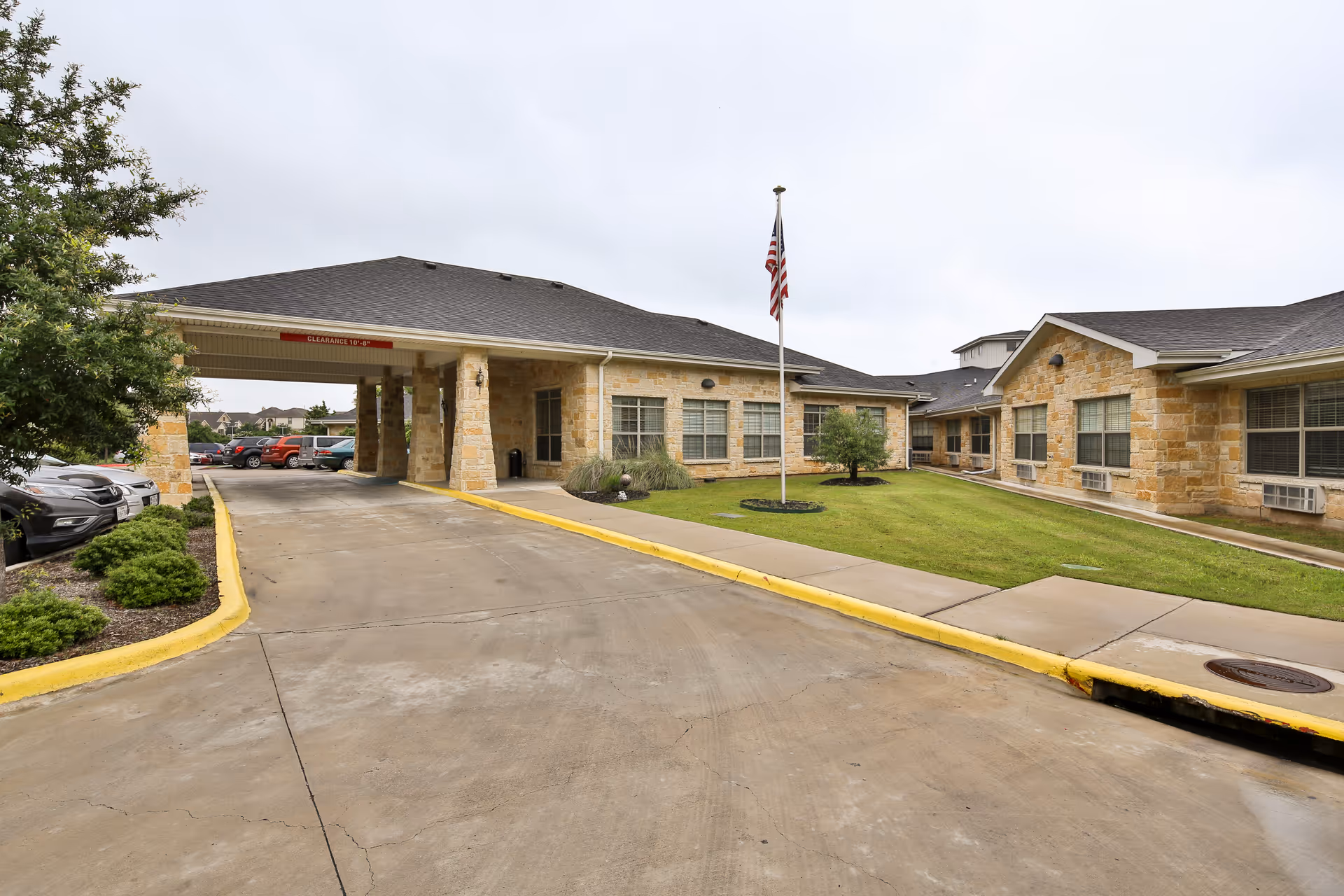 Exterior view of Southpark Meadows Nursing and Rehabilitation Center showing a covered driveway entrance with stone pillars, a flagpole with an American flag, green lawn, and parked cars to the left.