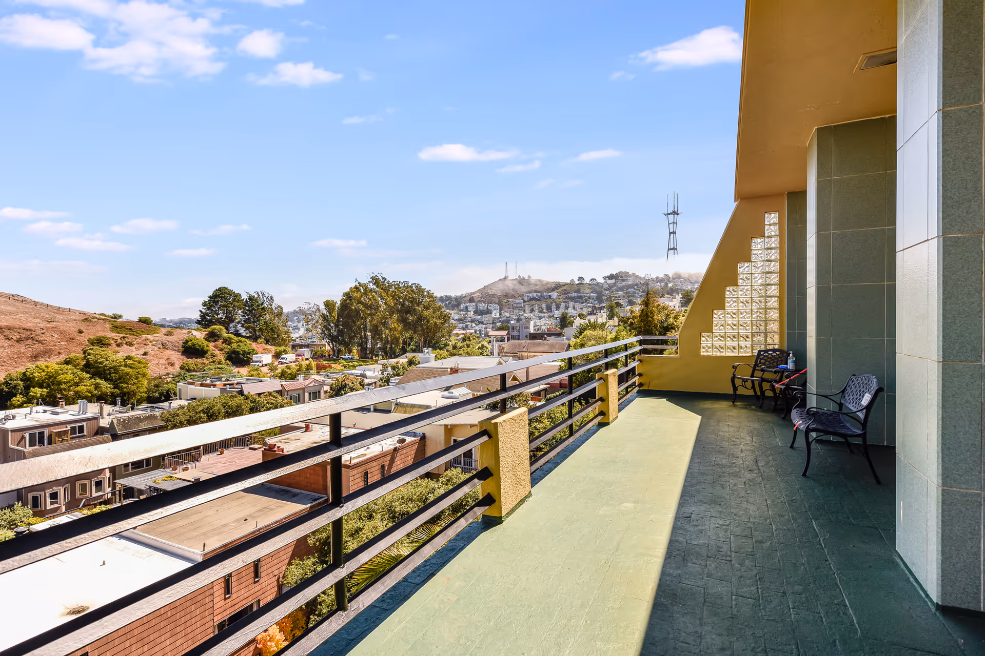 A spacious outdoor balcony with green flooring and black metal railings overlooking a residential neighborhood and hills in the distance under a blue sky with scattered clouds. There are two black metal benches and a small table on the balcony near the building wall.