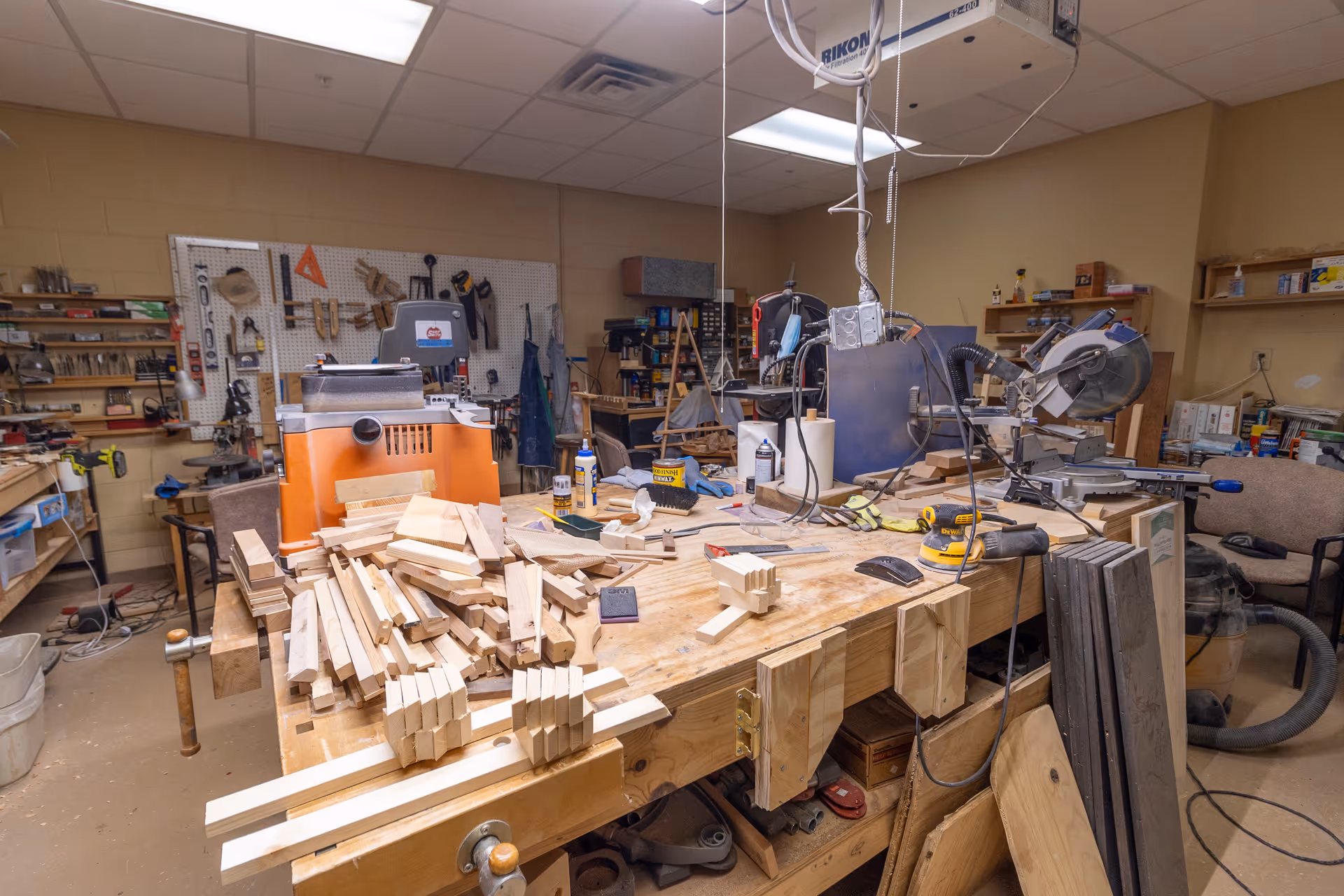 A woodworking workshop with a large workbench covered in various pieces of cut wood, tools, and equipment. The room has shelves filled with tools and supplies, pegboards with hanging tools, and woodworking machines including a miter saw and a planer. The ceiling has fluorescent lighting and an air filtration system.