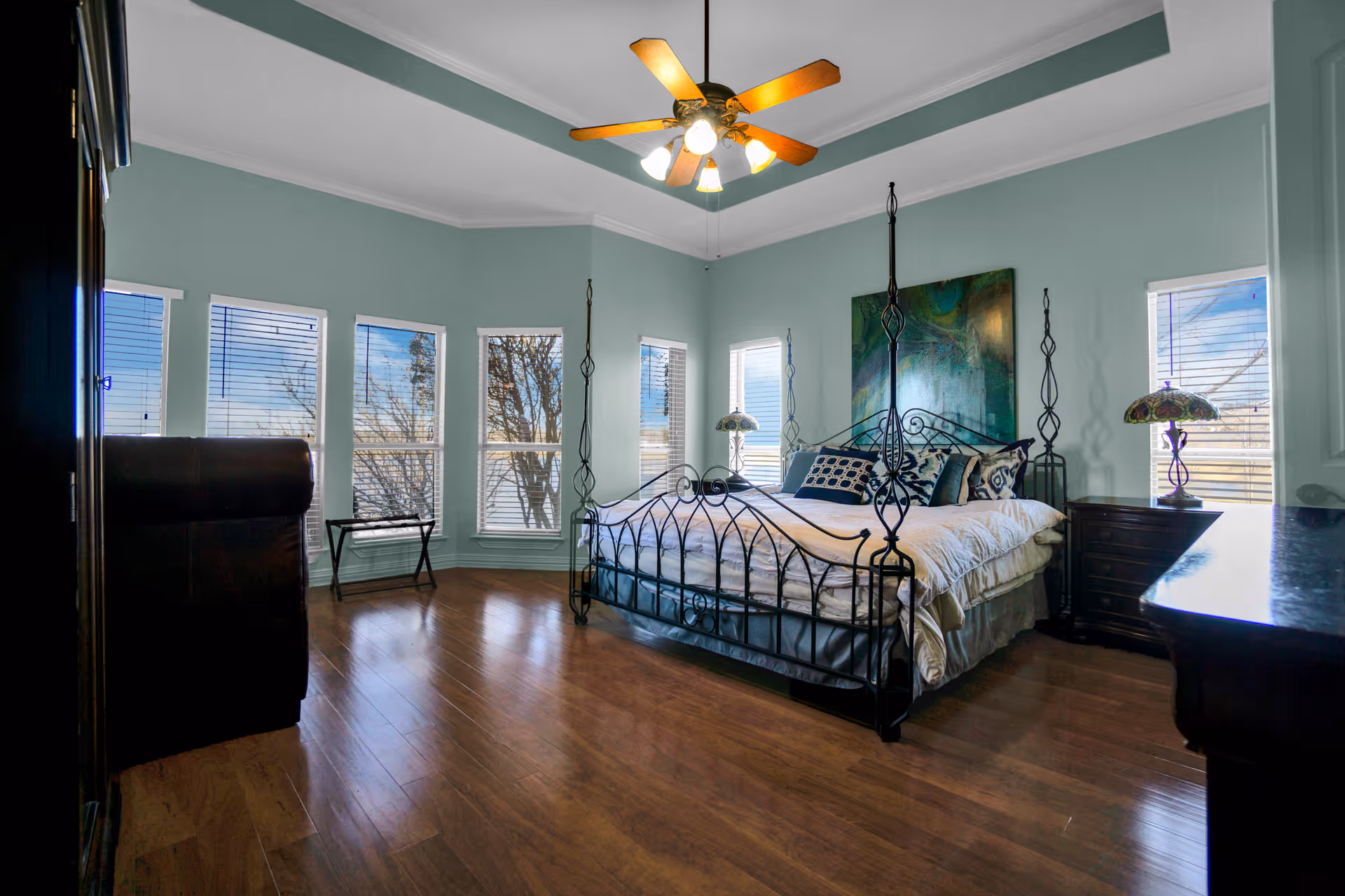 A spacious bedroom with light blue walls and wooden flooring. The room features a large black metal four-poster bed with white bedding and multiple decorative pillows. There are five windows with white blinds allowing natural light to enter. A ceiling fan with lights is mounted on the ceiling. On either side of the bed are dark wooden nightstands with decorative lamps. A large piece of abstract artwork hangs above the bed.