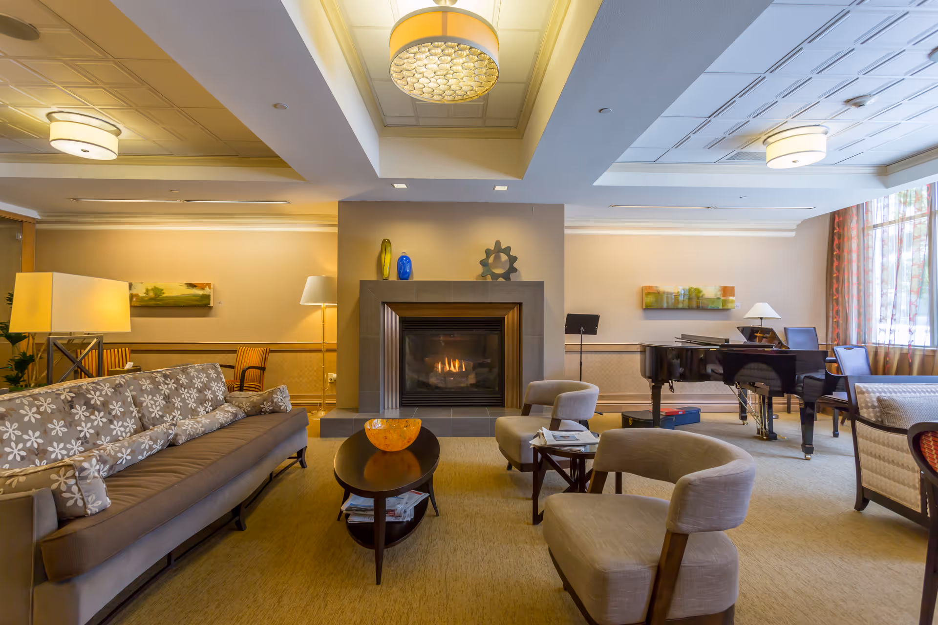 A cozy living room area with a lit fireplace in the center, surrounded by a floral-patterned sofa, two beige armchairs, a wooden coffee table with a decorative bowl, and a grand piano near a window with sheer curtains. The room is warmly lit with ceiling lights and lamps, and decorated with modern art pieces on the walls.