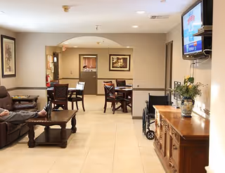 Interior view of a common area in a senior living facility with a brown leather couch, wooden coffee table, several wooden dining tables and chairs, a wheelchair, a wooden sideboard with a vase of flowers, and a wall-mounted television.