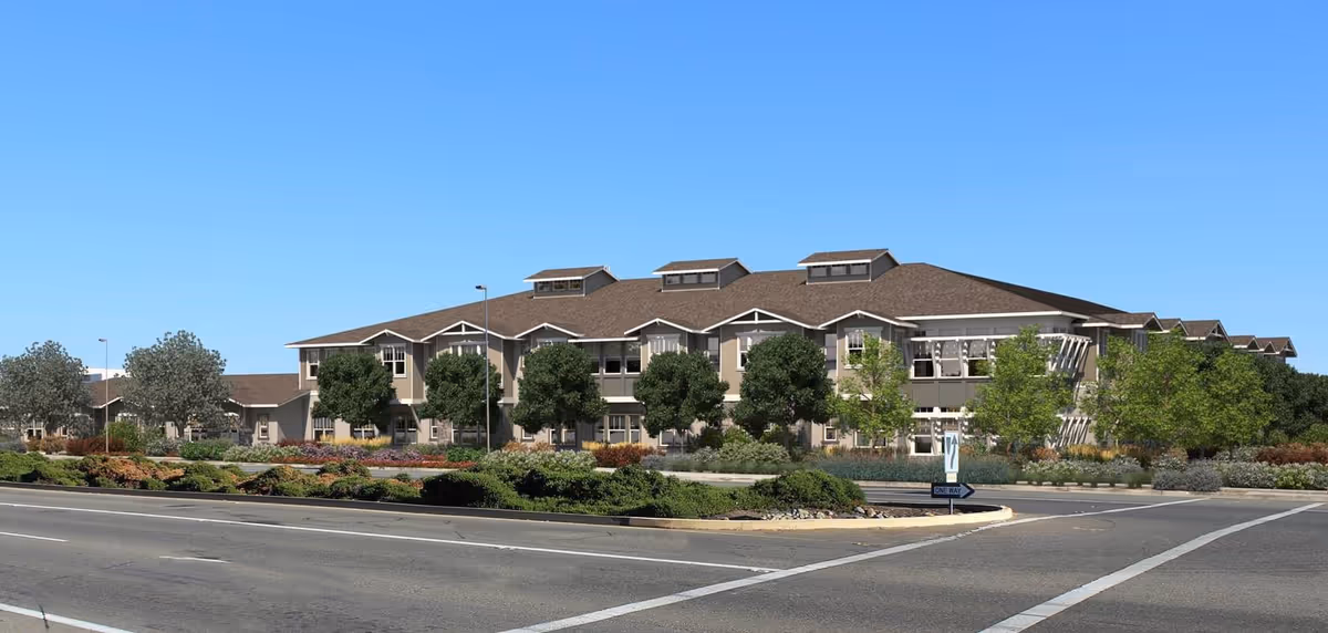 Exterior view of a large, two-story residential building with a brown roof and beige walls, surrounded by trees and landscaped greenery, under a clear blue sky.