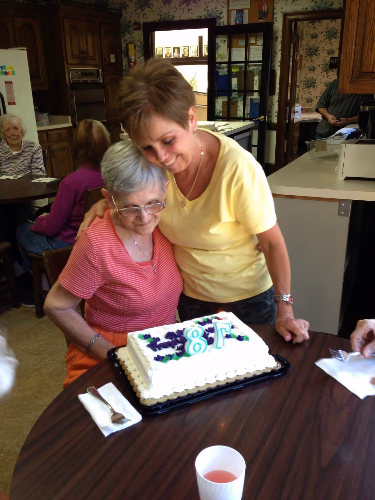 A woman embraces a seated woman as they lean over a rectangular birthday cake on a table in a communal dining area.
