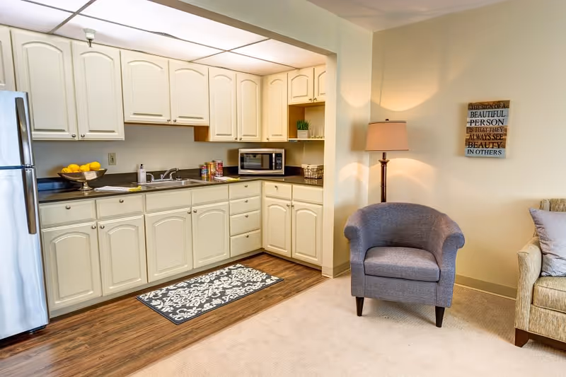 A small kitchen area with cream-colored cabinets, a stainless steel refrigerator, a microwave, and a countertop with a sink and some canned goods. Adjacent to the kitchen is a cozy living space with a gray upholstered armchair, a floor lamp, and a beige couch with pillows. A decorative wall sign is hung above the armchair.