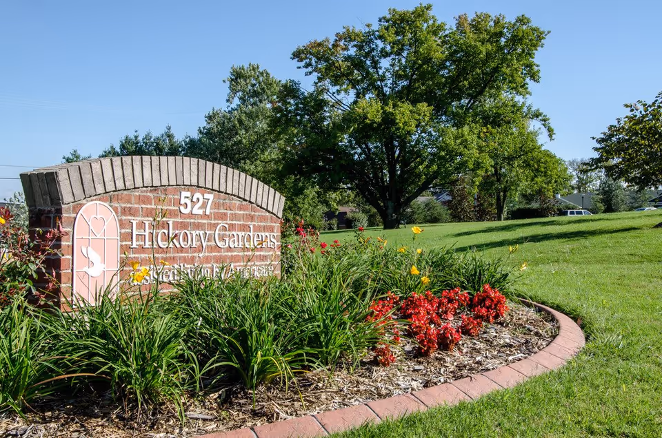A brick sign for Hickory Gardens assisted living facility surrounded by green grass, colorful flowers, and trees under a clear blue sky.