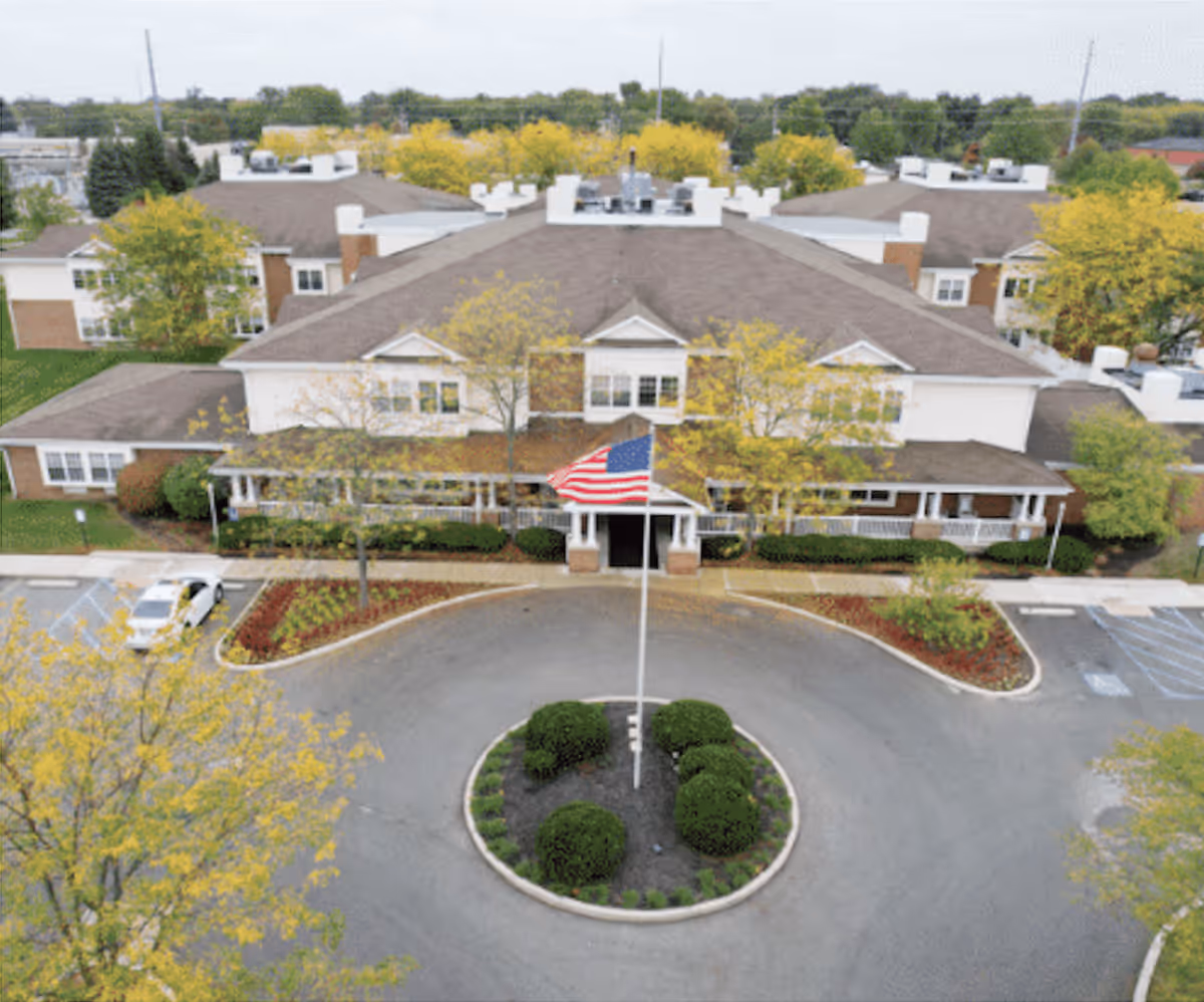 Aerial view of Carriage Court Senior Living facility showing a large building with a brown roof surrounded by trees with yellow leaves. In front of the building is a circular driveway with a landscaped island in the center featuring bushes and an American flag on a flagpole.