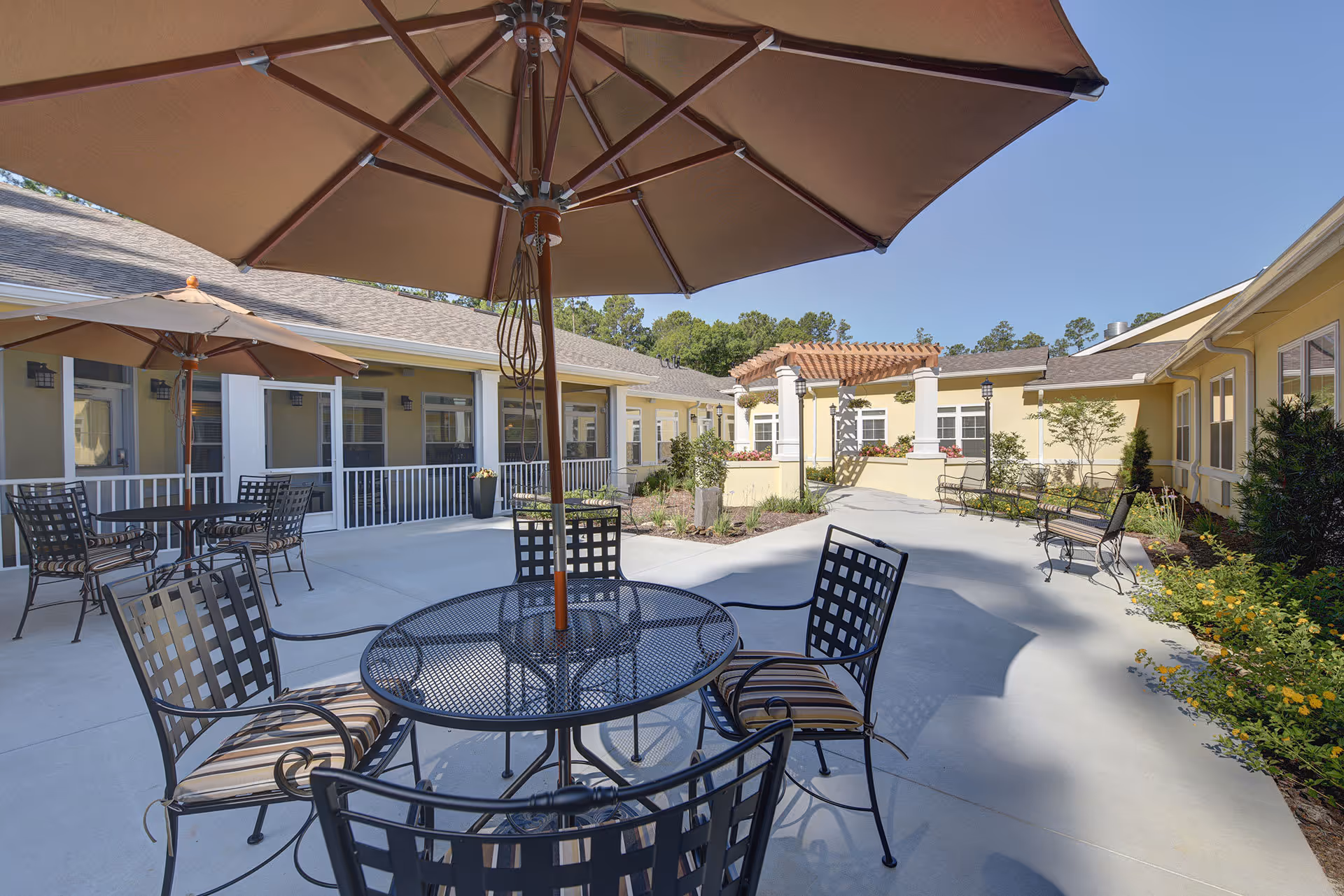 Outdoor courtyard area at The Addison of Bluffton featuring metal tables and chairs with striped cushions, large beige umbrellas providing shade, surrounded by yellow buildings with white trim, greenery, and a pergola in the background under a clear blue sky.