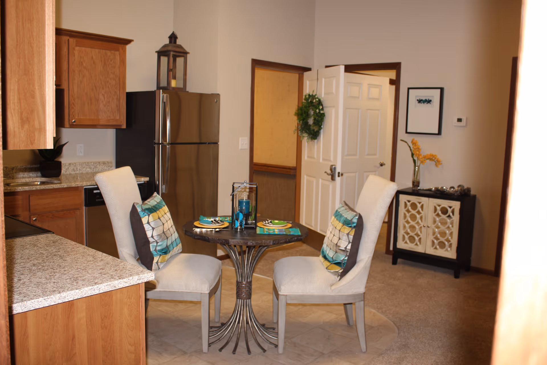 A small dining area in a kitchen with a round table set for two, featuring two cushioned chairs with patterned pillows. The kitchen has wooden cabinets, granite countertops, and a stainless steel refrigerator. In the background, there is a white door with a green wreath, a small cabinet with decorative items, and a framed picture on the wall.