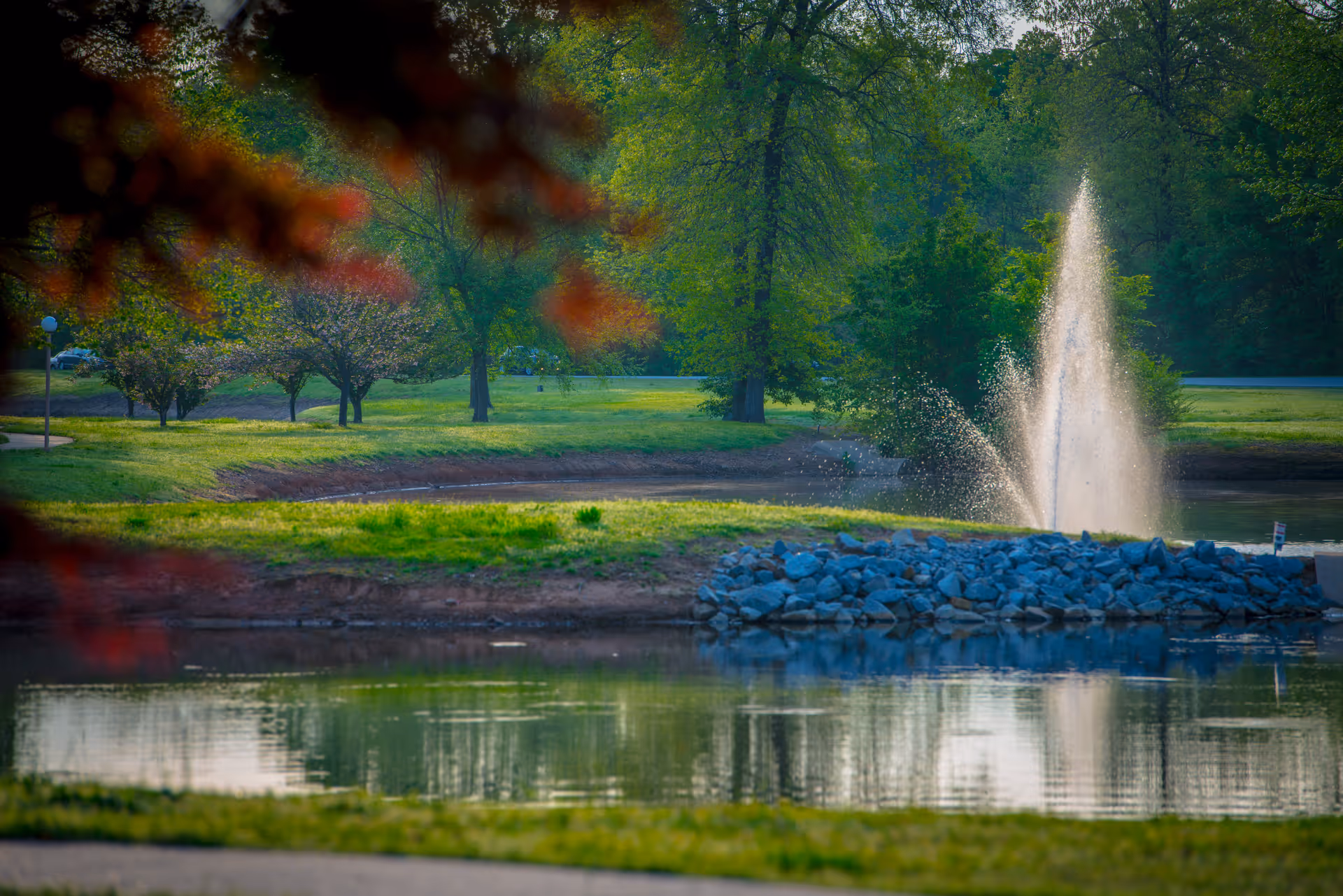 A serene outdoor scene at Parkway Village featuring a pond with a water fountain spraying water upwards. The pond is surrounded by green grass, trees, and a rocky embankment. The background shows a lush, wooded area with various trees and a clear sky.
