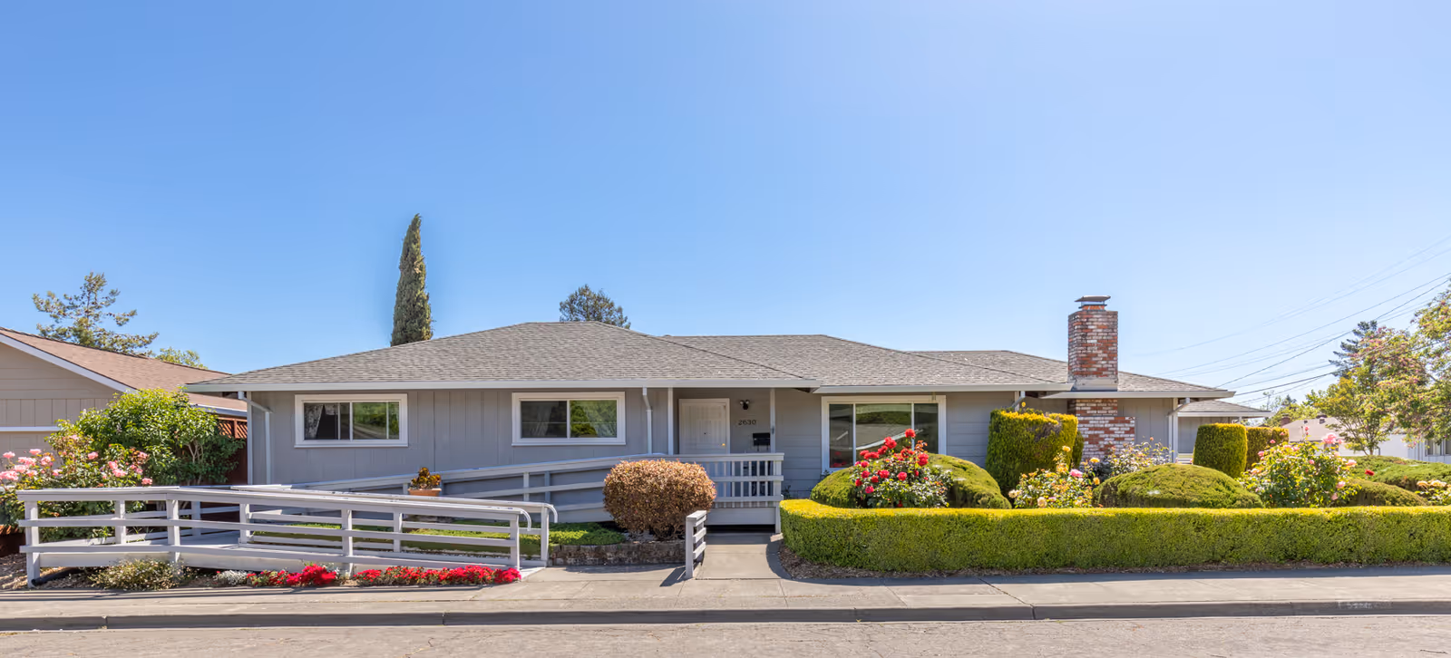 Single-story residential building with a gray exterior, a sloped roof, and a brick chimney. The front yard features neatly trimmed bushes, colorful flowers, and a wheelchair-accessible ramp leading to the front door. The sky is clear and blue.