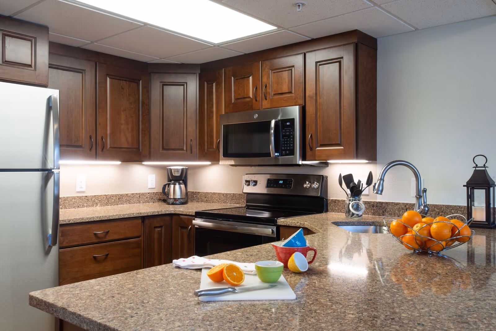 A modern kitchen with dark wooden cabinets, a stainless steel refrigerator, microwave, and stove. The countertop is granite with a sink and a bowl of oranges. There are colorful bowls and a cutting board with a sliced orange on the counter.
