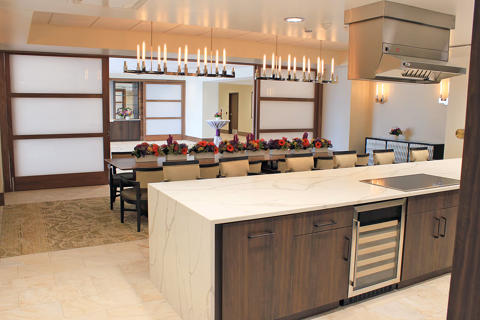 Modern kitchen and dining area with a large marble island countertop featuring a built-in wine cooler and stovetop. The dining table is decorated with colorful flower arrangements and surrounded by beige cushioned chairs. The room has wooden sliding doors with frosted glass panels, elegant chandeliers, and wall sconces providing warm lighting.