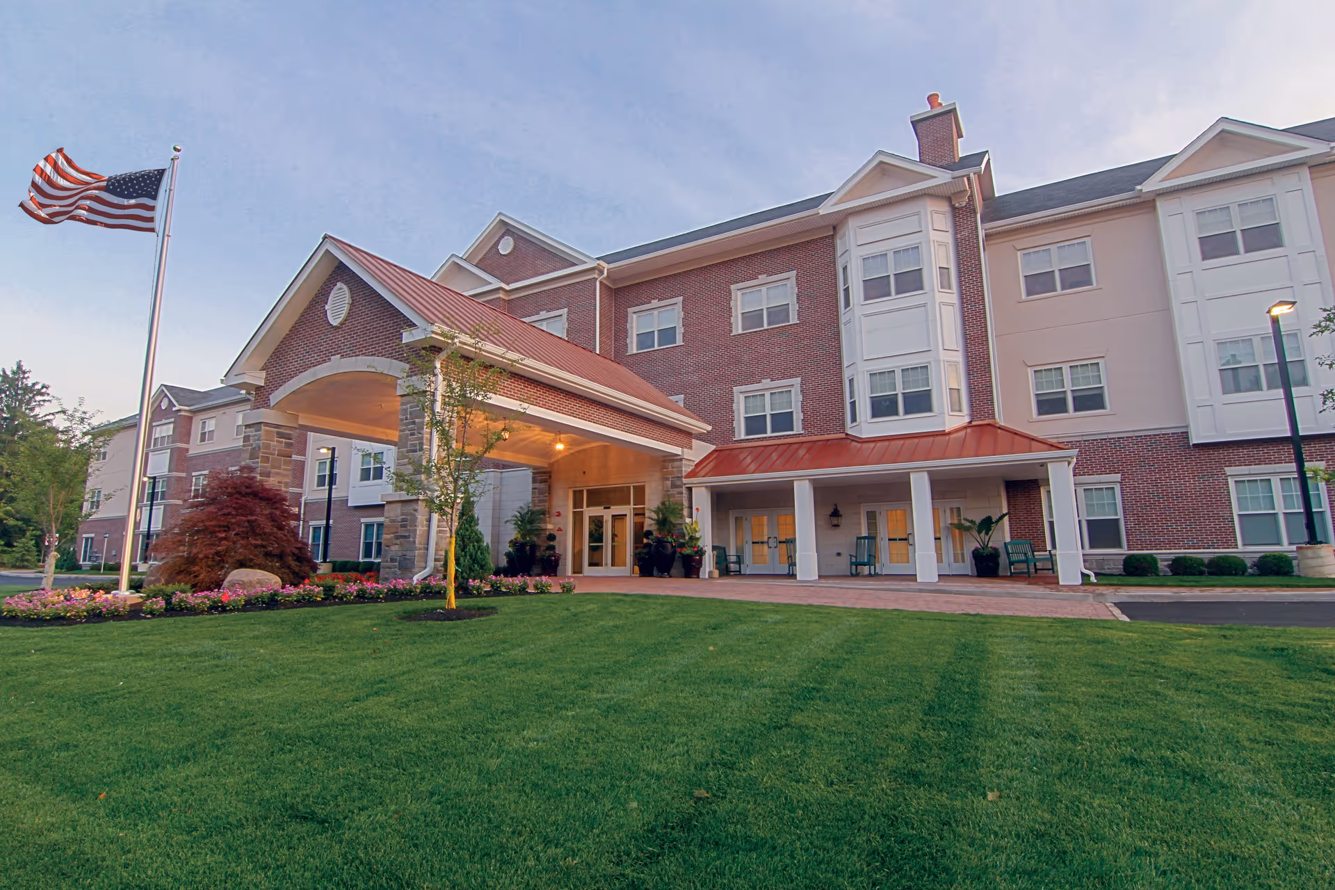 Front exterior view of a senior living facility building with a covered entrance, well-maintained green lawn, flower beds, and an American flag on a flagpole waving in the breeze under a clear sky.