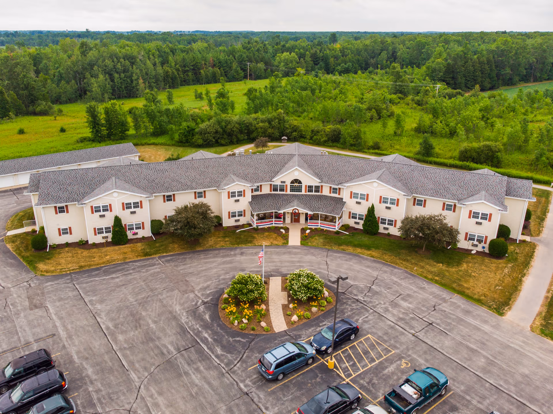 Aerial view of a large two-story senior living facility building named The Cottages at Meadowlands Senior Living, surrounded by green trees and grass. The building has a gray roof, beige walls, and red shutters. There is a circular driveway with a landscaped island in front, and several parked cars in the parking lot.