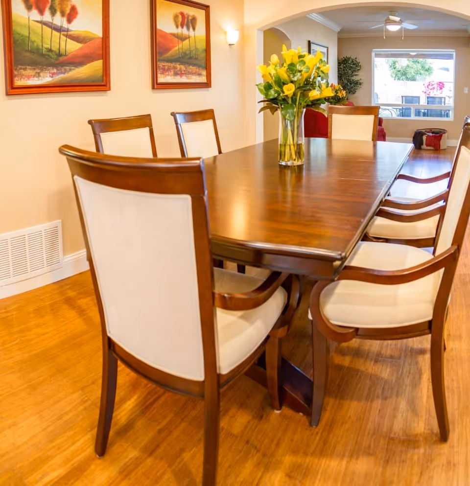 A dining room with a wooden table surrounded by six cushioned chairs. A vase with yellow flowers is placed in the center of the table. The room has wooden flooring, two framed landscape paintings on the wall, and a large window showing an outdoor patio area with chairs and greenery.