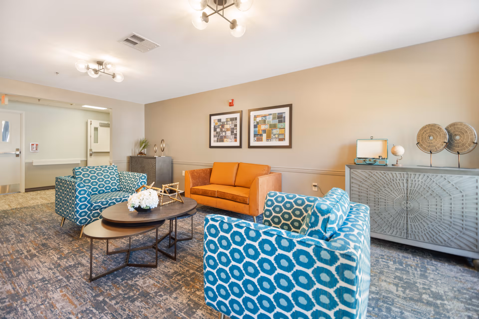 A bright and modern living room area with two blue patterned armchairs and an orange sofa arranged around two nested oval coffee tables. The tables hold a decorative white flower arrangement and a gold geometric sculpture. The room features beige walls with two framed abstract artworks, a gray sideboard with decorative items including a globe and circular sculptures, and carpeted flooring with a patterned design. Ceiling lights provide illumination.