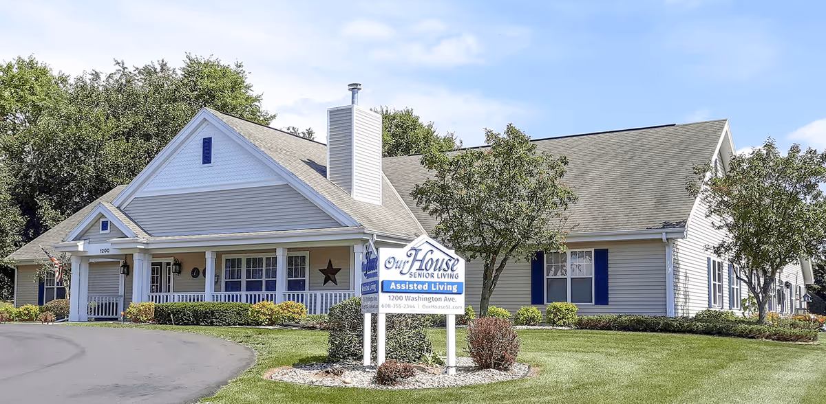 Exterior view of a single-story assisted living facility building with a light gray roof and beige siding. The building has a covered porch with white railings and several windows with blue shutters. There is a sign in front of the building that reads 'Our House Senior Living Assisted Living 1200 Washington Ave.' The surrounding area includes a paved driveway, green lawn, bushes, and trees under a clear blue sky.
