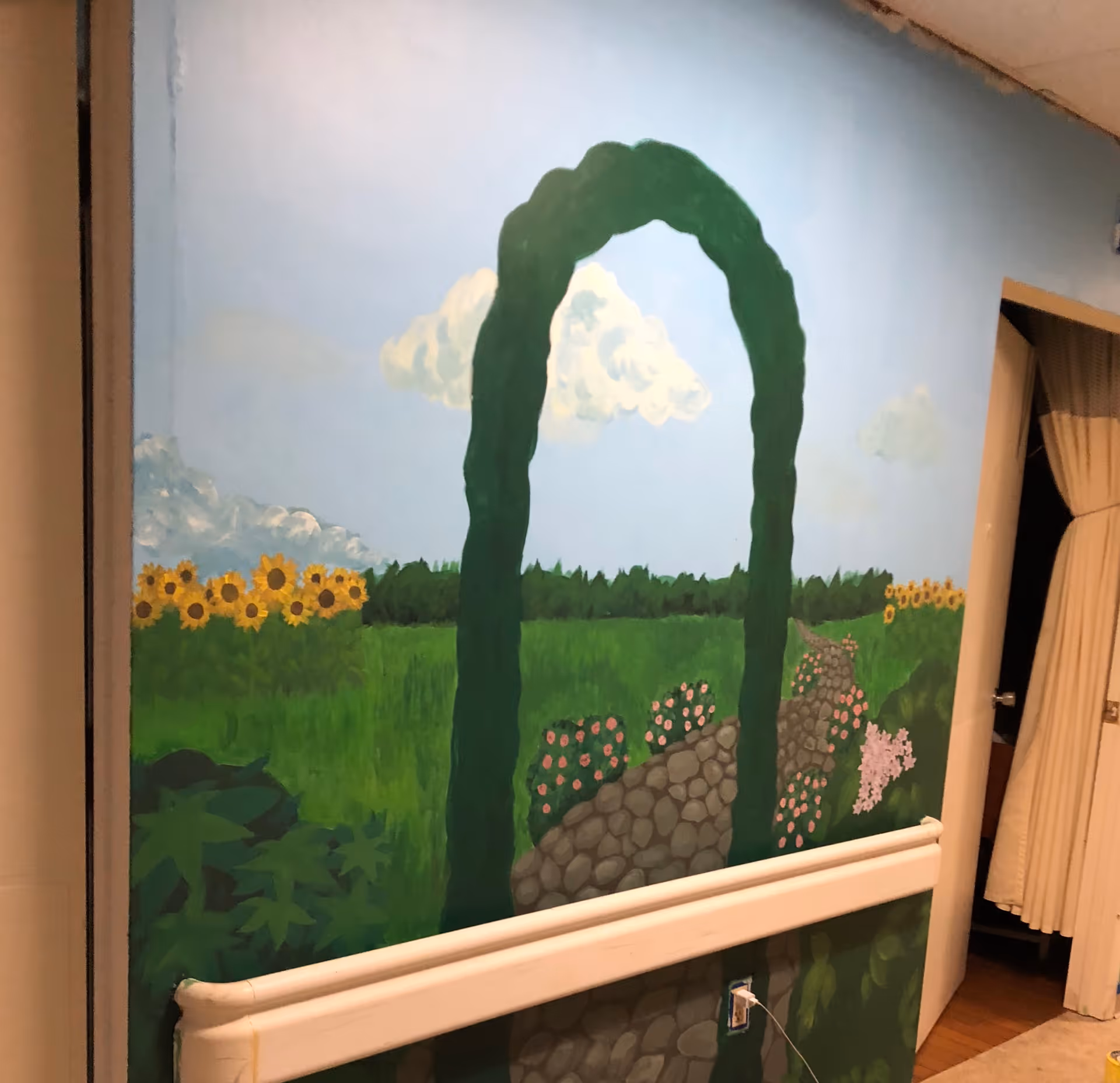 A painted mural on an interior wall depicting a garden scene with a green archway, a stone path, sunflowers, bushes with pink flowers, and a blue sky with clouds. There is a handrail attached to the wall below the mural and a partially open door with beige curtains on the right side.