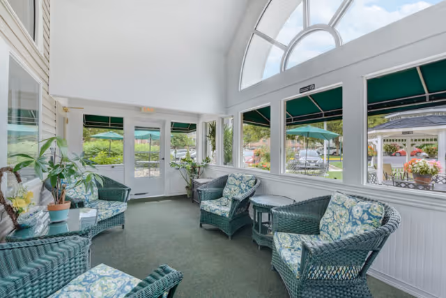 A bright sunroom with large windows and a high arched ceiling, furnished with green wicker chairs and sofas with floral cushions, small tables, and potted plants. Outside the windows, green umbrellas and a garden area are visible.