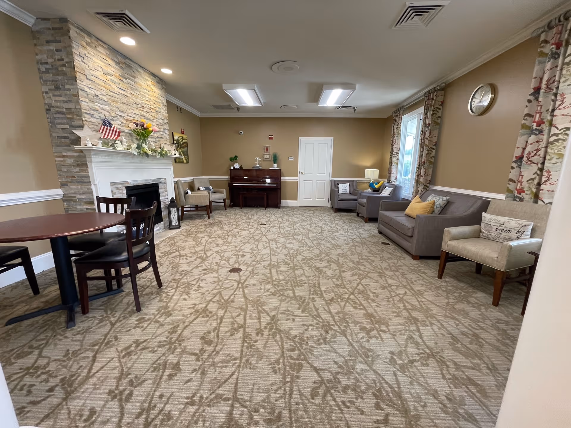 A spacious living room with beige walls and patterned carpet. The room features a stone fireplace with a white mantle decorated with flowers and an American flag. There are several chairs and sofas arranged along the walls, a round wooden table with chairs, a piano against the far wall, and large windows with floral curtains letting in natural light. A clock is mounted on the wall above one of the sofas.
