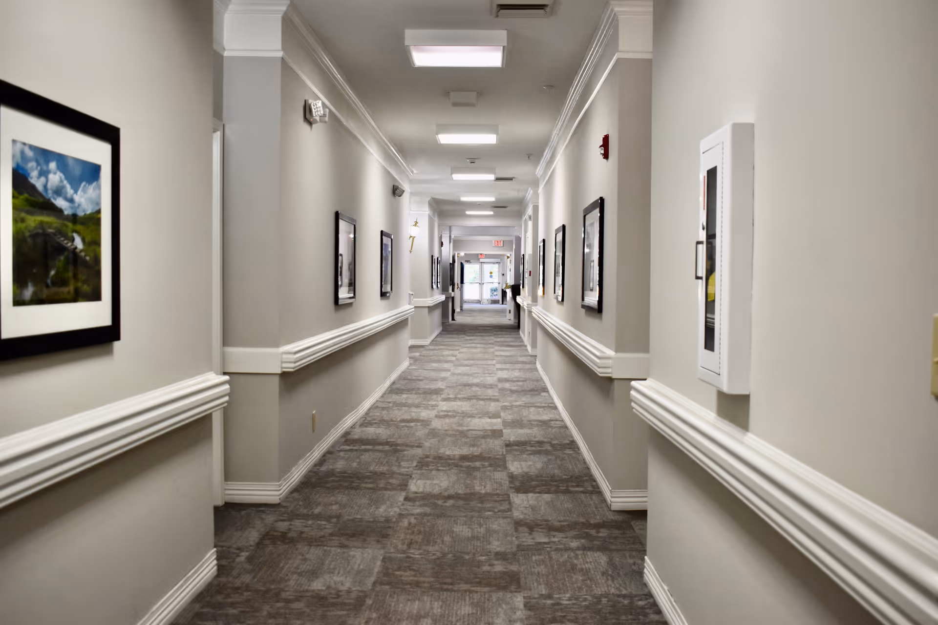 A long, well-lit hallway in a senior living facility with beige walls, carpeted floor, white trim, and framed pictures hanging on both sides. There is an exit door visible at the far end of the corridor.