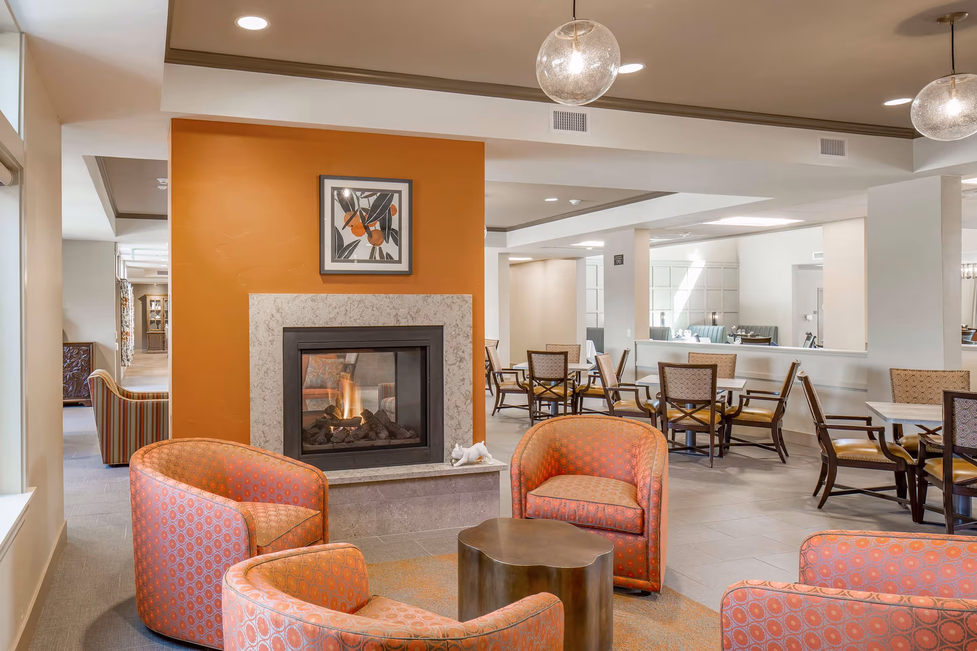 A cozy common area in Bruceville Point featuring a modern fireplace set in an orange accent wall with a framed abstract artwork above it. In front of the fireplace are four orange patterned armchairs arranged around a small, uniquely shaped coffee table. In the background, there are several dining tables with chairs, and the space is well-lit with ceiling lights and large windows.