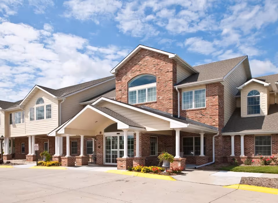 Exterior view of a two-story assisted living facility with a brick and beige siding facade, large windows, and a covered entrance with white columns. The sky is partly cloudy.