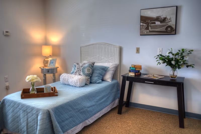 A neatly made single bed with a light blue quilt and multiple decorative pillows in a cozy room. A small round side table with a lamp and a framed photo is beside the bed. A wooden tray with a vase of white flowers and a mug is placed on the bed. Across from the bed is a black table with a stack of books, an open magazine, and a vase with white flowers. A black and white vintage photo hangs on the wall above the table.