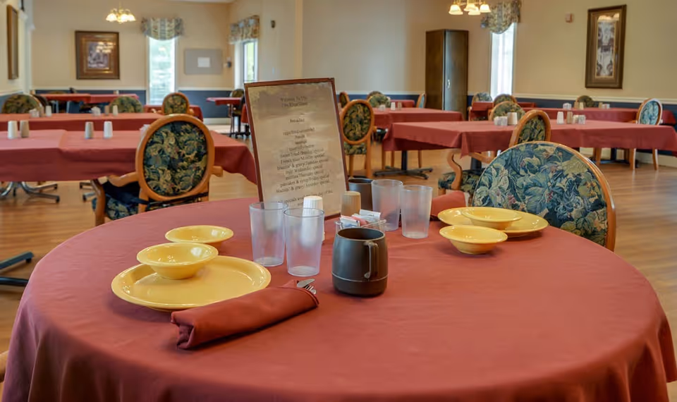 A dining room in Rock Point Nursing Center with round tables covered in red tablecloths. Each table is set with yellow plates and bowls, glasses, a coffee mug, and a menu card. The chairs have floral patterned upholstery. The room has wooden flooring, framed artwork on the walls, and windows letting in natural light.