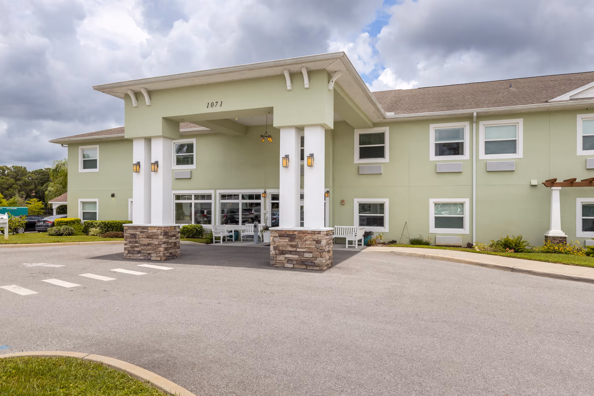 Front exterior view of a two-story senior living facility building with a covered entrance supported by white columns with stone bases. The building is painted light green with white trim around the windows. There are benches near the entrance and a driveway in front. The sky is cloudy.