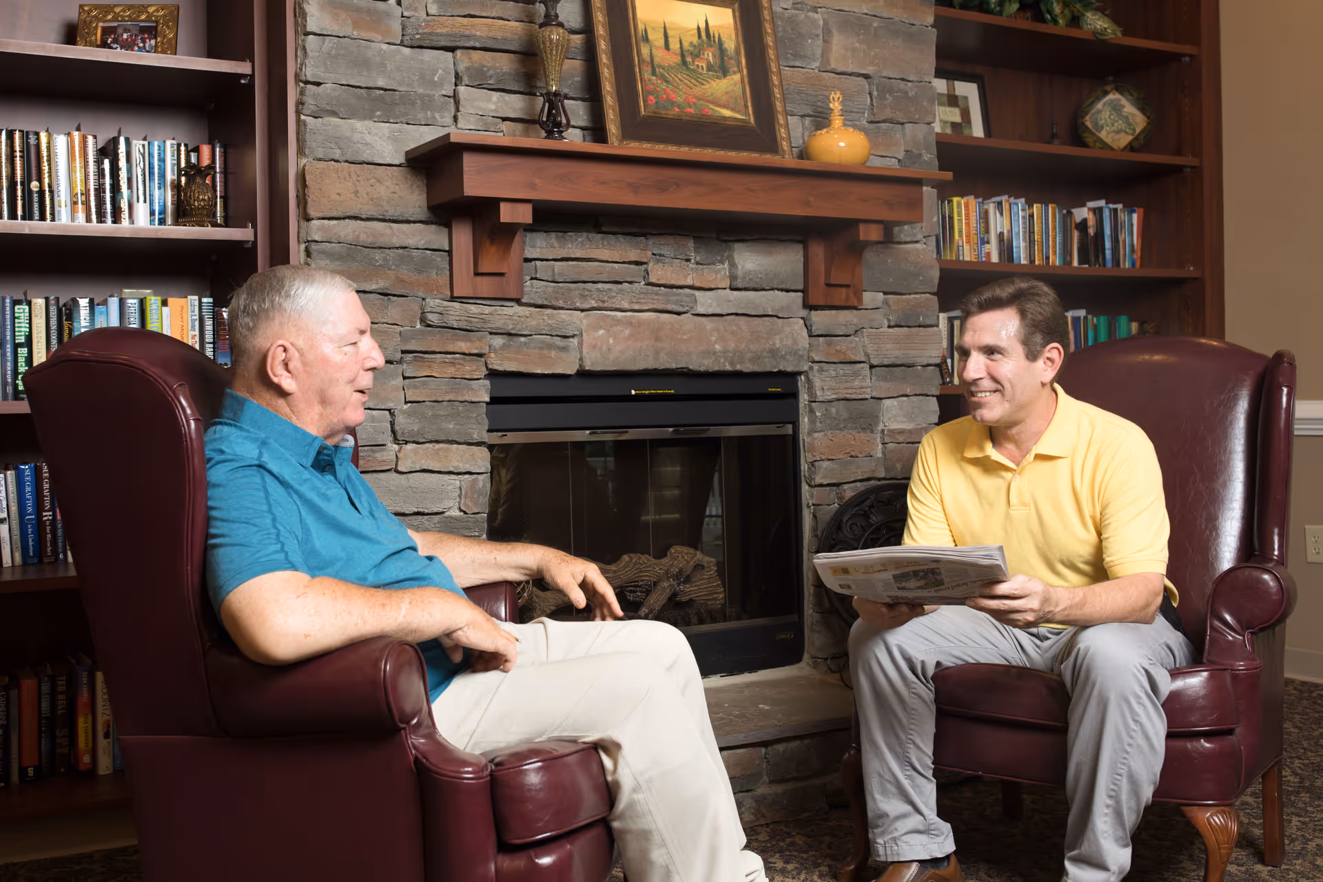 Two men sitting in leather armchairs facing each other in a cozy room with a stone fireplace and wooden bookshelves filled with books. One man is wearing a blue shirt and light pants, and the other is wearing a yellow polo shirt and light pants, holding a newspaper and smiling.