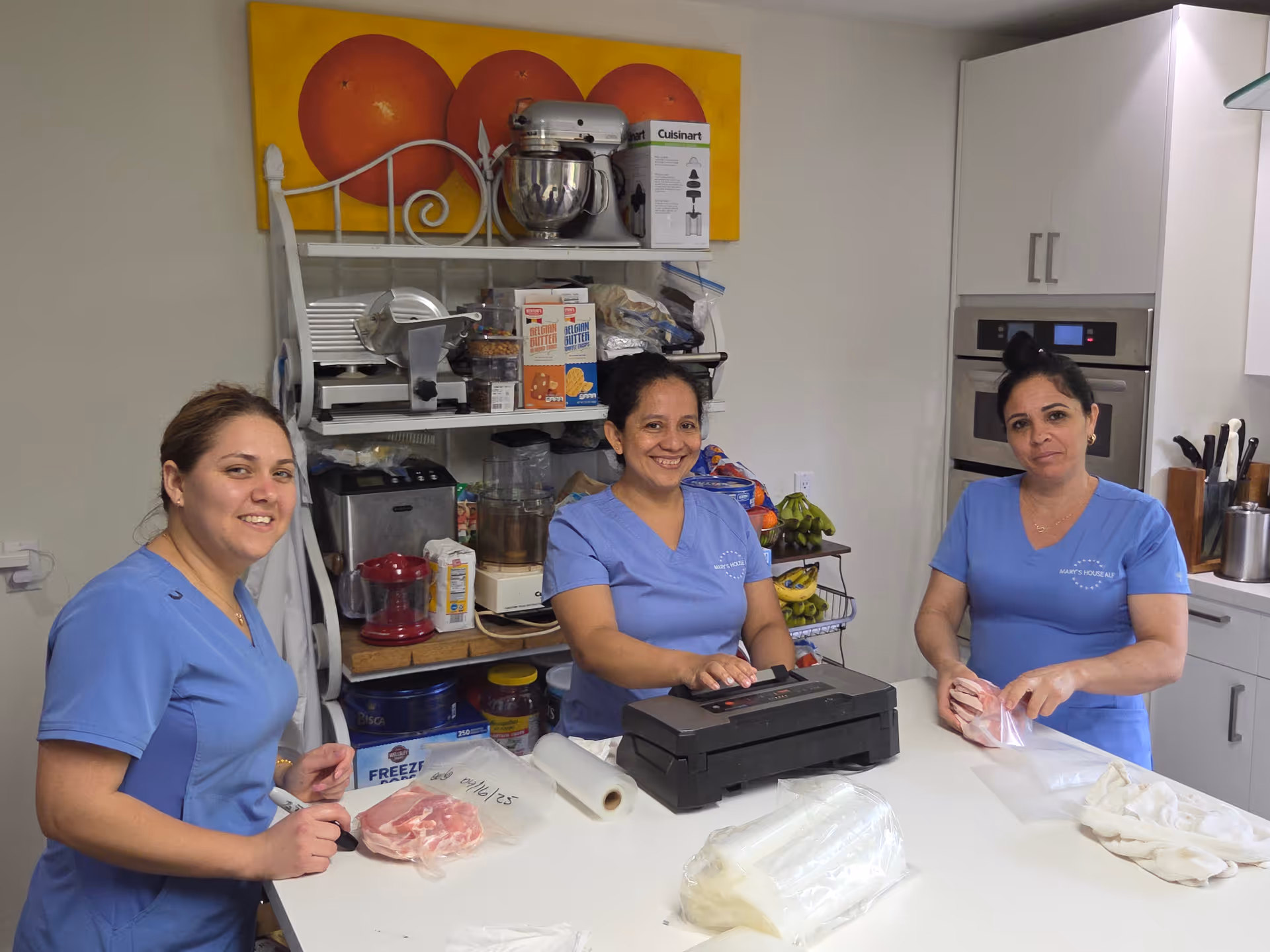 Three women in blue uniforms standing around a kitchen counter, packaging raw meat into plastic bags. Behind them is a kitchen shelf with various kitchen appliances and food items, and a wall painting of three oranges.