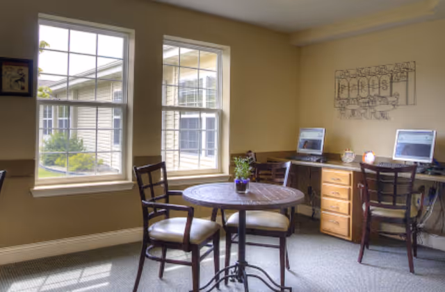 A bright room with two large windows showing an exterior view of another building. Inside, there is a round wooden table with three chairs around it and a small potted plant on top. Against the wall, there is a wooden desk with two computers and two chairs. The walls are painted beige, and there is a decorative metal wall art above the desk.