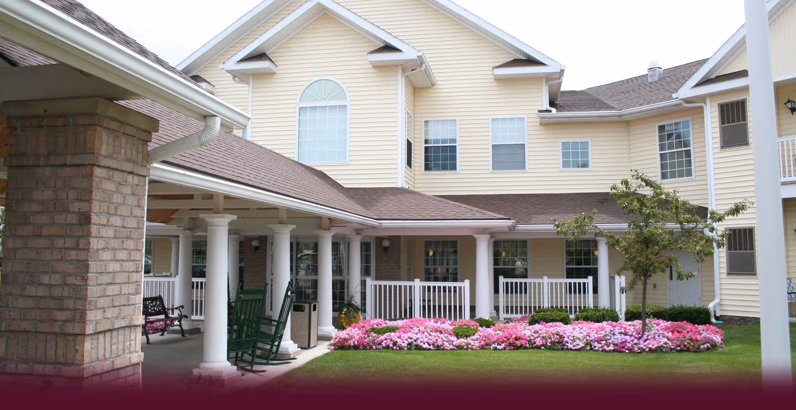 Exterior view of a senior living community building with beige siding, white columns, and a covered porch area featuring rocking chairs and benches. There is a well-maintained garden with pink and white flowers and a small tree in front of the porch.