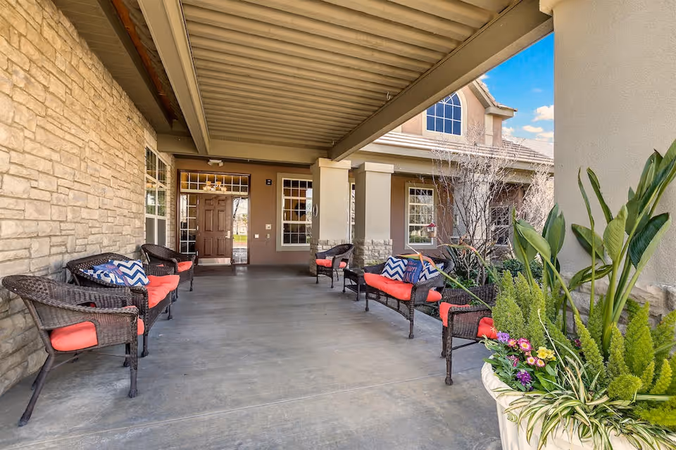 Covered front porch with wicker chairs and red cushions, potted plants, and the entrance to the senior living facility.