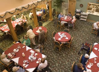 A dining room in a senior living facility with several round and rectangular tables covered with red tablecloths. Elderly residents are seated at the tables, eating and conversing. The room has carpeted floors with a floral pattern, wooden chairs, and decorative garlands hanging along the upper wall.