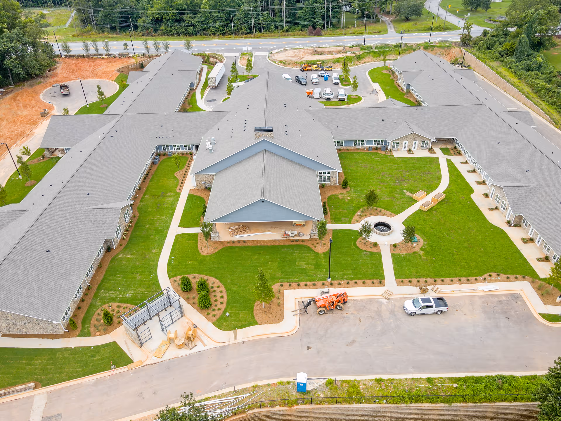 Aerial view of Manor Lake Hamilton Mill assisted living and memory care facility showing a large building complex with multiple wings surrounding a central courtyard with green lawns, pathways, and a circular fire pit area. Several vehicles and construction equipment are parked near the building, and the surrounding area includes roads and trees.