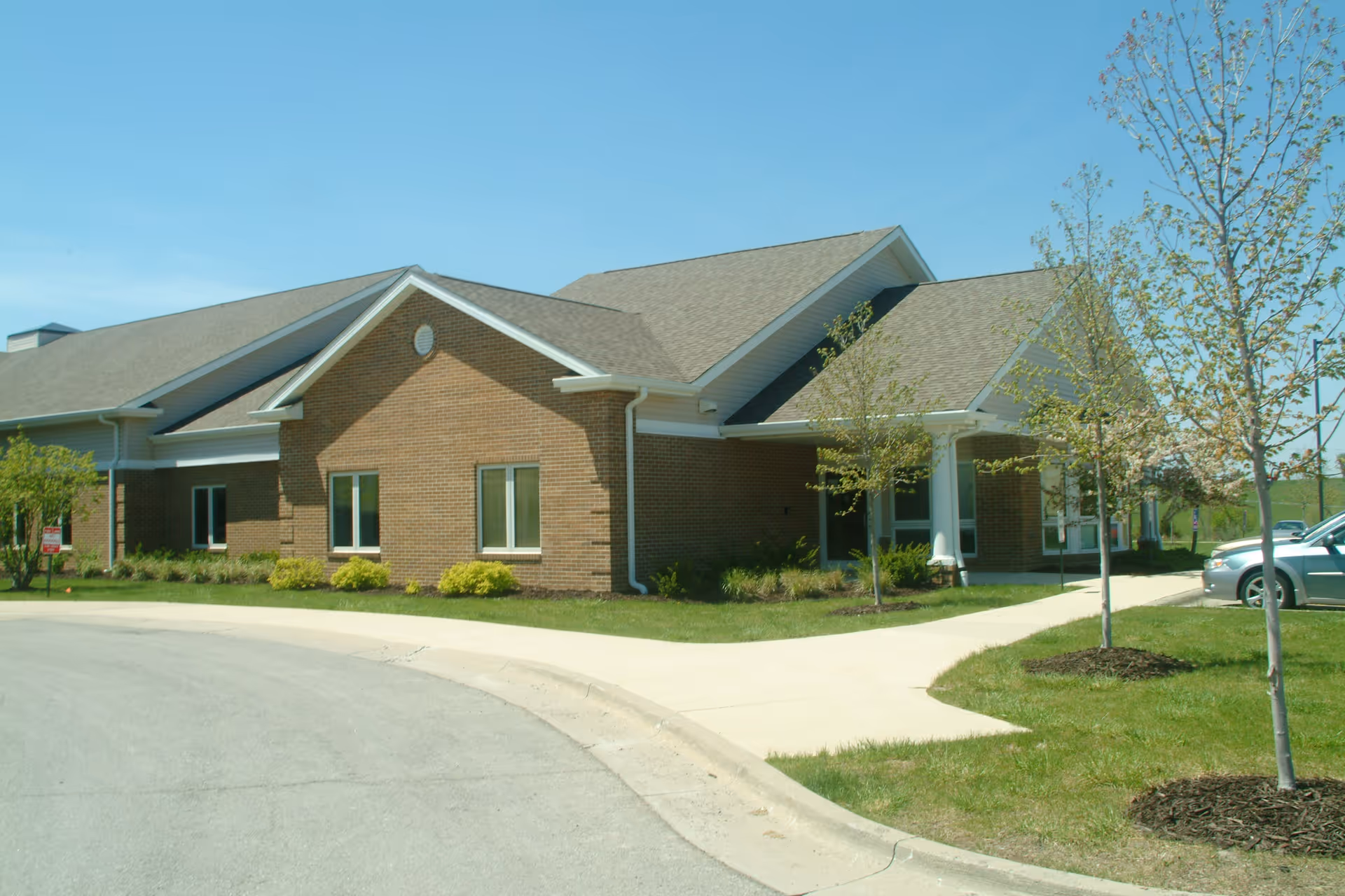 Exterior view of a single-story brick building with a gabled roof, surrounded by a sidewalk, small trees, and a parking area with cars under a clear blue sky.
