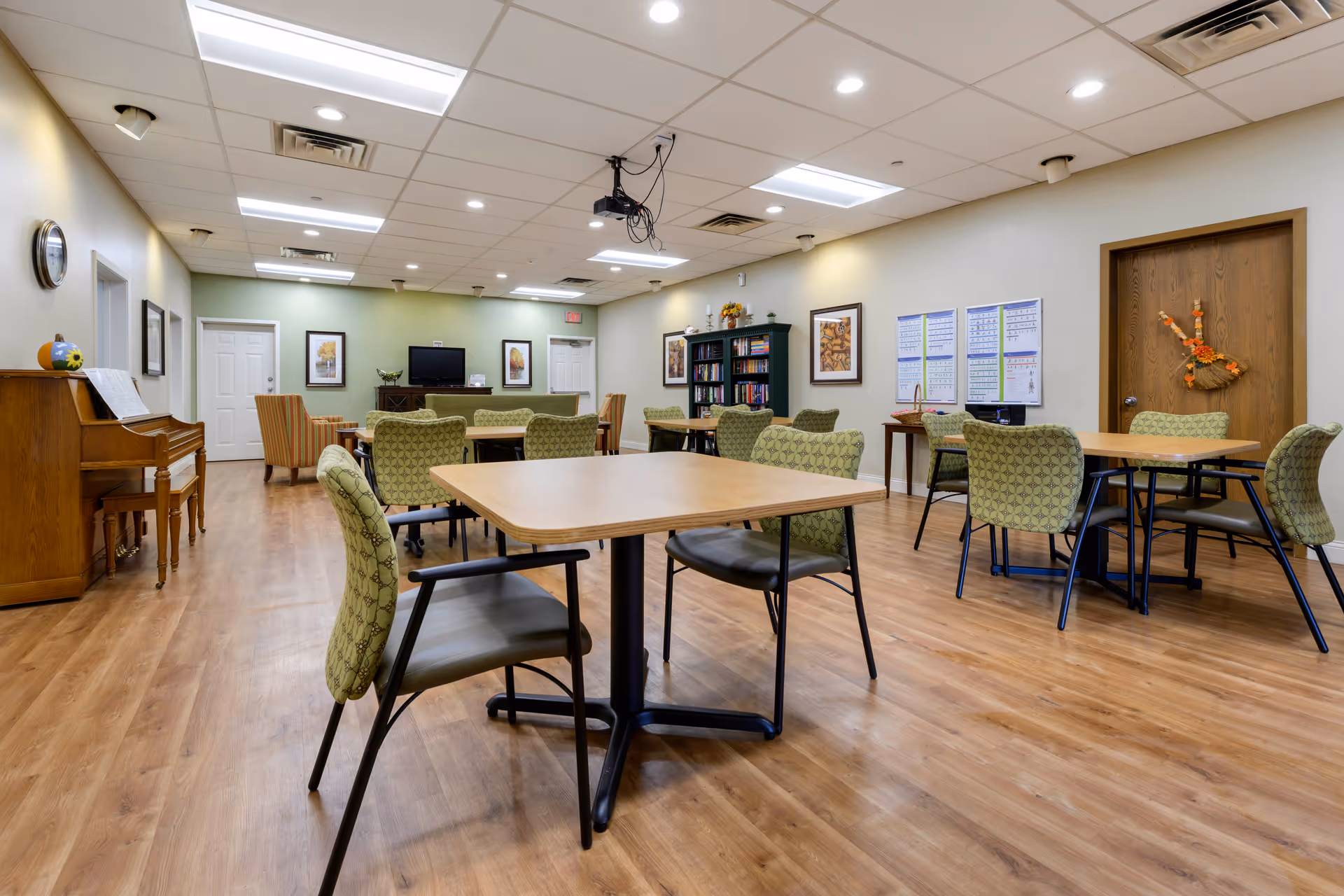 A spacious common area in a senior living facility with several tables and green patterned chairs arranged on a wooden floor. The room features a piano on the left side, a TV mounted on the far wall, bookshelves, framed artwork, and a door decorated with a fall-themed wreath. The ceiling has recessed lighting and a projector mounted in the center.