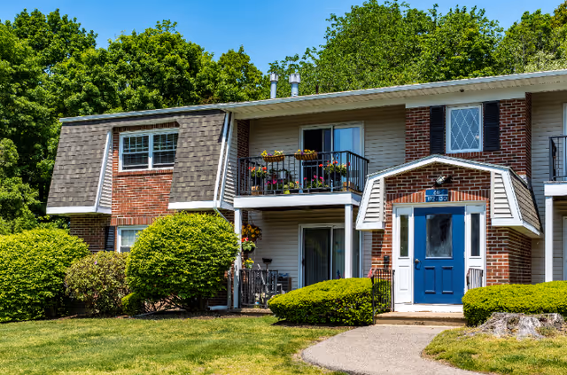 Exterior view of a two-story residential building with brick and siding facade, featuring a blue front door, balconies with flower pots, and surrounded by green bushes and trees under a clear blue sky.