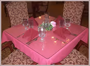 A square dining table covered with a pink tablecloth set for four people. Each place setting includes a folded pink napkin, a fork, and a water glass. There is a small green plant centerpiece and a candle in the middle of the table. The chairs around the table have patterned upholstery.