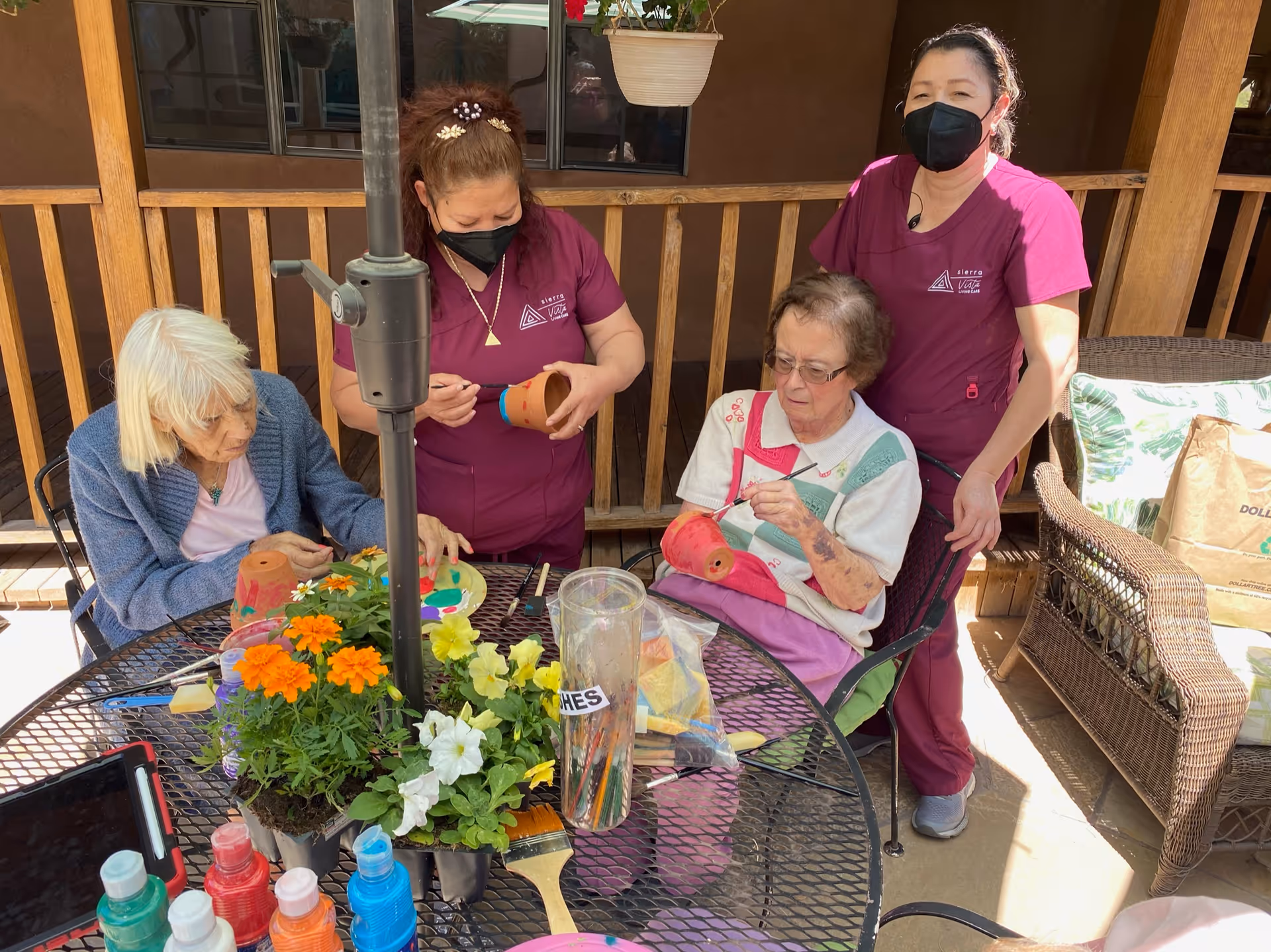 Two elderly women sitting at a round outdoor table painting small flower pots, accompanied by two caregivers wearing maroon uniforms and black face masks. The table has various painting supplies, colorful flowers, and a clear container labeled 'BRUSHES'. The setting appears to be a sunny patio with wooden railings and wicker furniture.