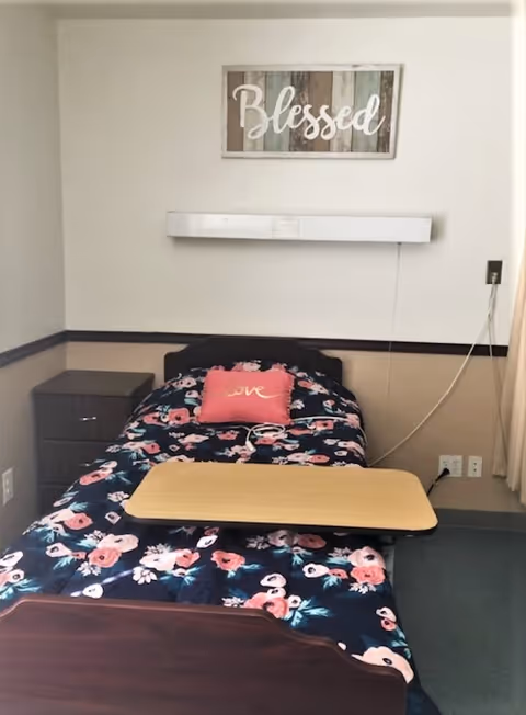 A single bed with a floral patterned comforter and a pink pillow with the word 'love' on it. Above the bed is a wall decoration with the word 'Blessed'. There is a wooden bedside table with drawers to the left of the bed and a beige overbed table positioned over the bed. The room has beige and white walls with a dark trim and a window with curtains on the right side.