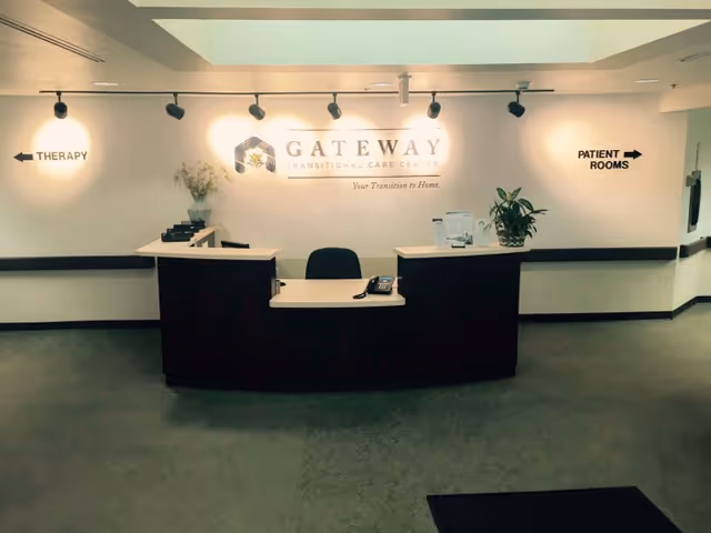 Reception desk area at Gateway Transitional Care Center with a sign on the wall behind the desk displaying the facility name and slogan. There are directional signs on the wall indicating therapy to the left and patient rooms to the right. The desk has a phone, some papers, and a plant on it.