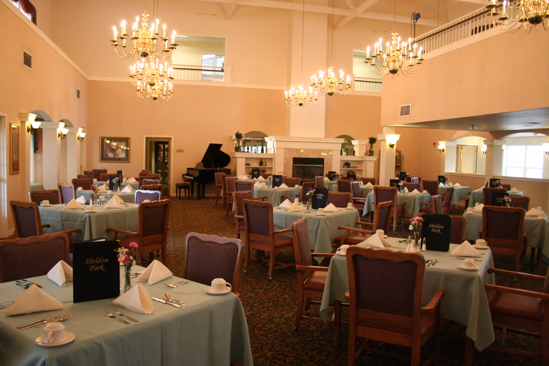 A spacious dining room with multiple tables covered in light blue tablecloths, each set with white napkins, cups, and silverware. The room features elegant chandeliers hanging from a high ceiling, a grand piano in the back corner, and a fireplace with decorative plants on the mantel. The chairs are wooden with purple cushions, and menus labeled 'Sheldon Park' are placed on several tables.