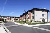 Exterior view of a multi-story senior living facility building with a curved driveway, landscaped grass areas, and clear blue sky.