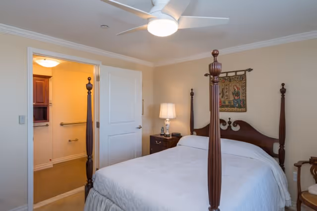 A bedroom with a four-poster wooden bed covered with a white bedspread. There is a nightstand with a lamp and a clock next to the bed. A tapestry hangs on the beige wall above the bed. The door to an adjacent bathroom is open, showing a grab bar and part of the bathroom interior. A ceiling fan with a light is mounted on the ceiling.