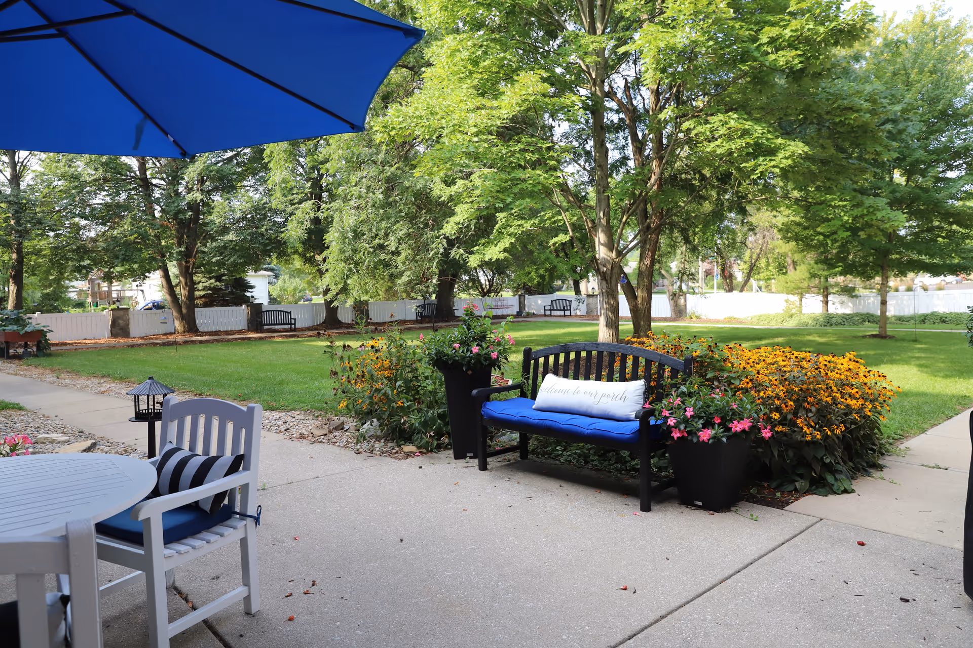 Patio seating with a blue-umbrella table and a bench with cushions beside flower beds overlooking a grassy yard and trees.